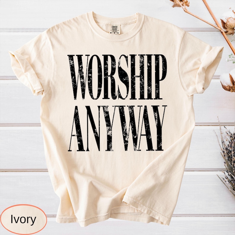 May include: Ivory-colored t-shirt with the words "WORSHIP ANYWAY" printed in bold, black letters. The shirt is made of a soft material and has rolled-up sleeves. The word "Ivory" is printed at the bottom of the shirt.