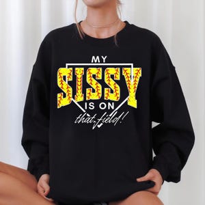 May include: Black sweatshirt with the text "MY SISSY IS ON that field!" in yellow and red. The word "SISSY" is designed to look like a softball. The sweatshirt is a crew neck style.