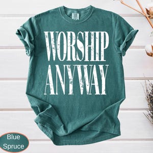 May include: A blue spruce t-shirt with the words "WORSHIP ANYWAY" printed in large white capital letters. The shirt has a crew neck and short sleeves. The shirt is folded and has a tag on the upper back.