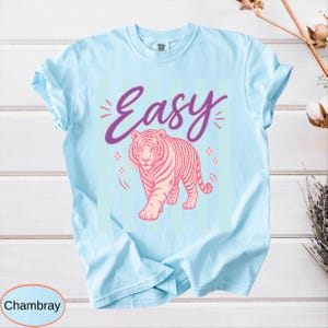 May include: Light blue t-shirt with a pink tiger illustration and the word "Easy" in purple script. The design includes star accents. The shirt is made of a soft, comfortable material.