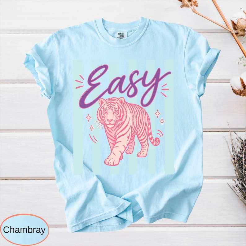 May include: Light blue t-shirt with a pink tiger illustration and the word "Easy" in purple script. The design includes star accents. The shirt is made of a soft, comfortable material.