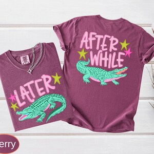 Puede incluir: Dos camisetas color bayas con dise&ntilde;os gr&aacute;ficos. Una camiseta presenta la palabra "LATER" con un caim&aacute;n verde y estrellas. La otra camiseta tiene "AFTER WHILE" con un caim&aacute;n verde y estrellas. Una cadena de plata est&aacute; en la primera camiseta.