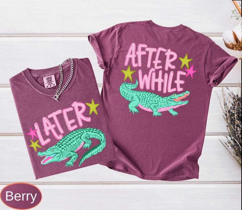 Puede incluir: Dos camisetas color bayas con dise&ntilde;os gr&aacute;ficos. Una camiseta presenta la palabra "LATER" con un caim&aacute;n verde y estrellas. La otra camiseta tiene "AFTER WHILE" con un caim&aacute;n verde y estrellas. Una cadena de plata est&aacute; en la primera camiseta.