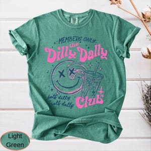 May include: Light green t-shirt with a graphic design. The design features a smiling face with crossed-out eyes, a drink, and the words "MEMBERS ONLY the Dilly Dally Club". The text "half dilly, half dally" is also included.