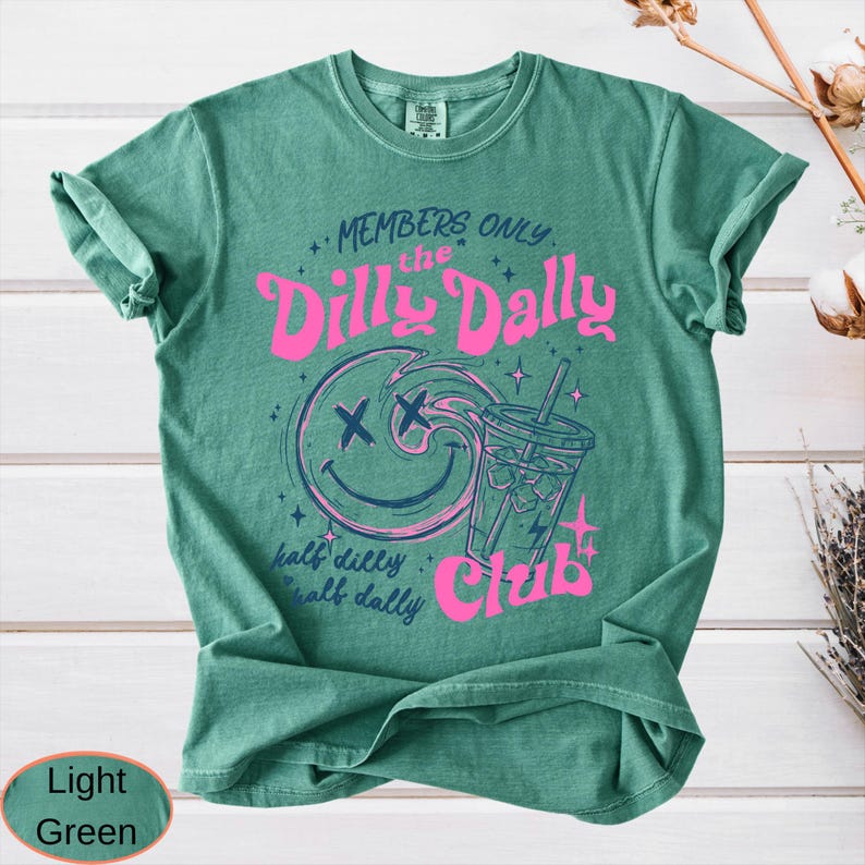 May include: Light green t-shirt with a graphic design. The design features a smiling face with crossed-out eyes, a drink, and the words "MEMBERS ONLY the Dilly Dally Club". The text "half dilly, half dally" is also included.