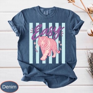 May include: Denim blue t-shirt featuring a pink tiger illustration with the word "Easy" in a cursive font. The design includes light blue vertical stripes and star accents. The shirt has rolled-up sleeves.