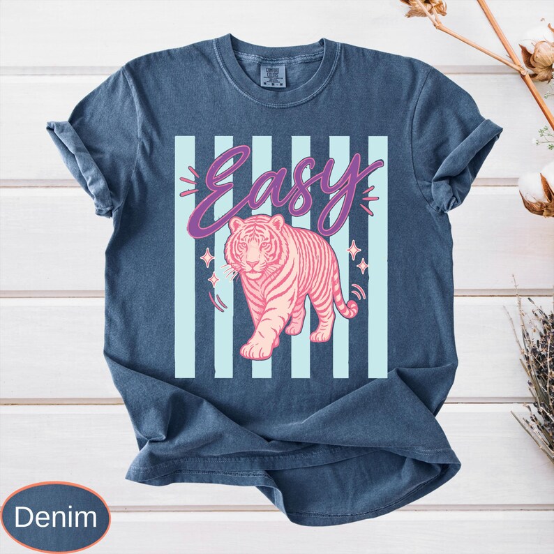 May include: Denim blue t-shirt featuring a pink tiger illustration with the word "Easy" in a cursive font. The design includes light blue vertical stripes and star accents. The shirt has rolled-up sleeves.