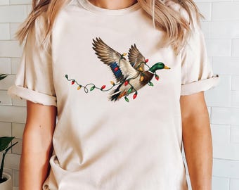 May include: A cream-colored short-sleeve t-shirt featuring a graphic of a mallard duck in flight, wrapped in a string of colorful red, green, and yellow Christmas lights. The duck has a green head, brown chest, and brown and white wings.