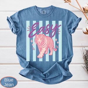 May include: Blue jean t-shirt with a pink tiger graphic and the word "Easy" in a script font. The tiger is walking forward, and the background has light blue vertical stripes. The shirt is made of a soft, comfortable material.