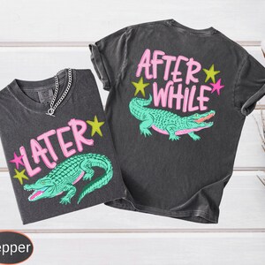 Puede incluir: Dos camisetas grises oscuras con gr&aacute;ficos de caimanes. Una dice "LATER" con estrellas rosas y amarillas, la otra dice "AFTER WHILE". Los caimanes son verdes con bocas rojas. Se ve un collar de cadena plateada.