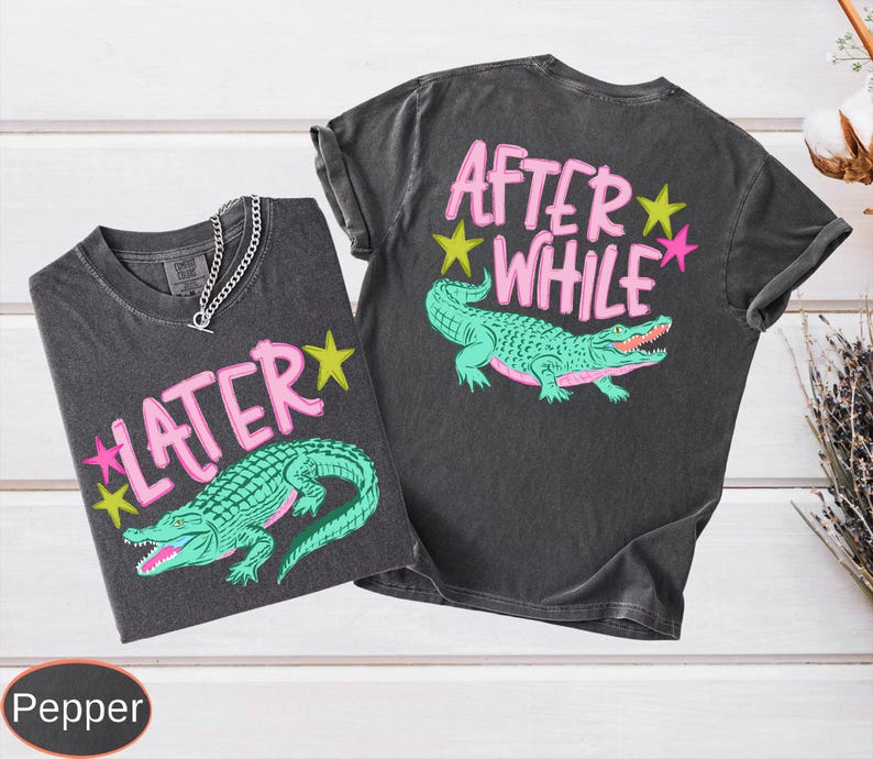 Puede incluir: Dos camisetas grises oscuras con gr&aacute;ficos de caimanes. Una dice "LATER" con estrellas rosas y amarillas, la otra dice "AFTER WHILE". Los caimanes son verdes con bocas rojas. Se ve un collar de cadena plateada.