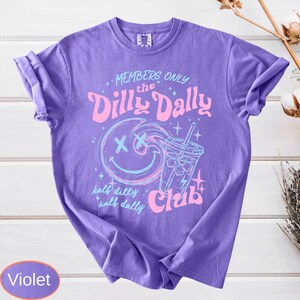 May include: Violet-colored t-shirt with the text "MEMBERS ONLY the Dilly Dally Club" in pink and blue. The design includes a smiley face and a drink with ice. The shirt is made of a soft material.