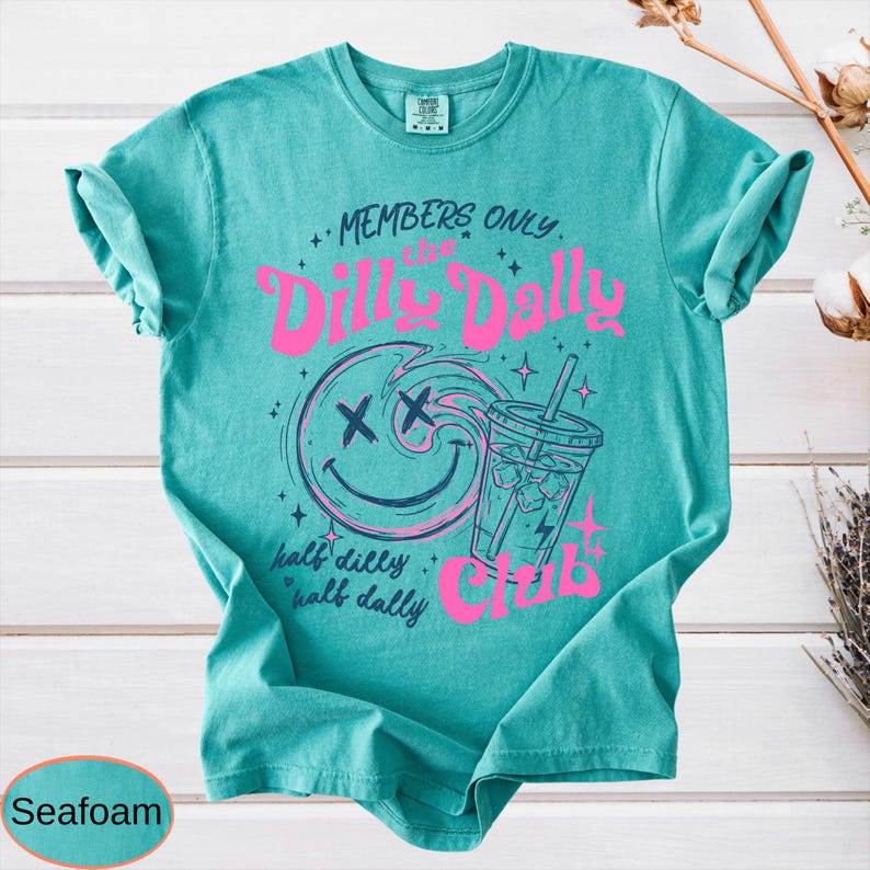 May include: Seafoam green t-shirt with a graphic design. The design features the text "MEMBERS ONLY" above "The Dilly Dally Club" in pink. A smiling face with an "X" for eyes and a drink with ice are also in the design.