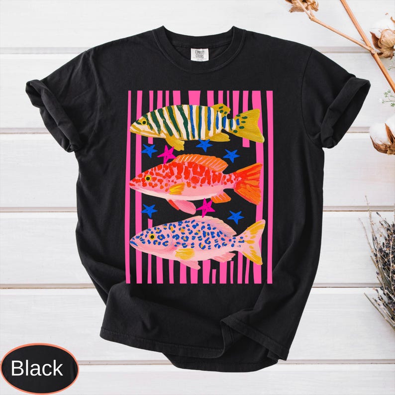 May include: Black t-shirt featuring a colorful graphic of three fish. The fish are set against a pink and white striped background with blue and pink star accents. The word "Black" is at the bottom.