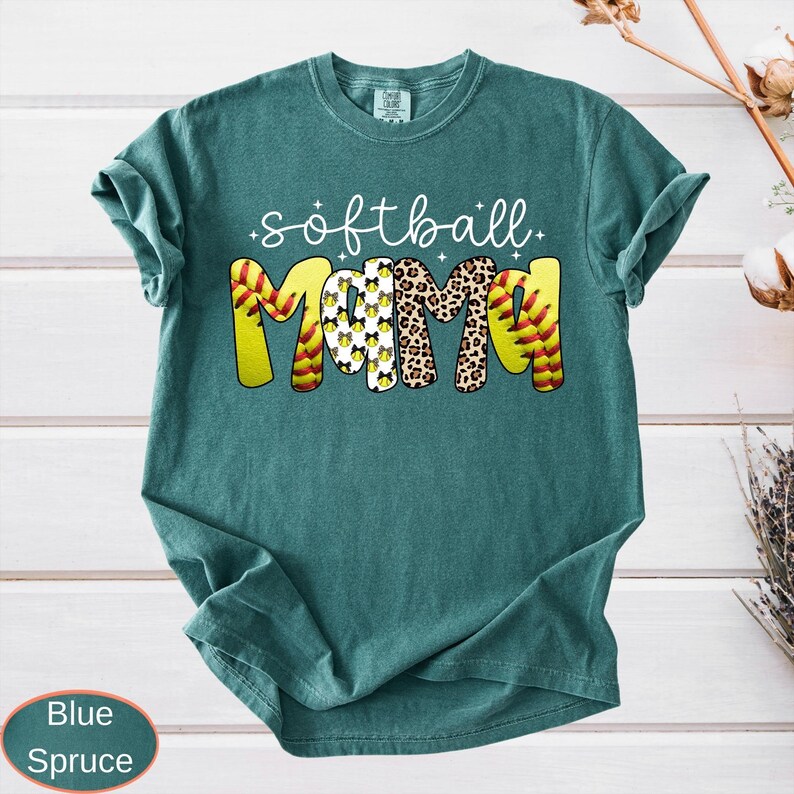 A vintage wash garment showing the text softball in cursive over the word MAMA. The thick letters showcase different designs like sports ball stitching, spotted animal print, and repeated bows, complete with tiny decorative sparkles.