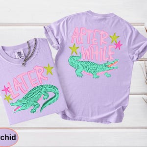 Puede incluir: Dos camisetas color orqu&iacute;dea con dise&ntilde;os a juego. Cada camiseta presenta un gr&aacute;fico de caim&aacute;n verde y el texto "LATER" o "AFTER WHILE" en rosa, con acentos de estrellas rosas y amarillas. Las camisetas se muestran sobre una superficie de madera blanca.