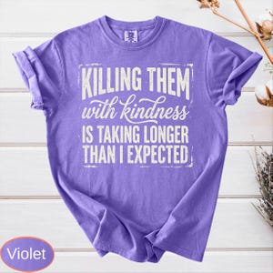 Op de afbeelding: Een violetkleurig T-shirt met korte mouwen en witte tekst die luidt: "KILLING THEM with kindness IS TAKING LONGER THAN I EXPECTED." Het shirt heeft een ronde hals en een relaxte pasvorm, met de afbeelding gecentreerd op de voorzijde.
