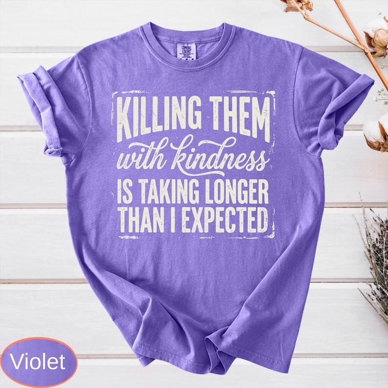 Op de afbeelding: Een violetkleurig T-shirt met korte mouwen en witte tekst die luidt: "KILLING THEM with kindness IS TAKING LONGER THAN I EXPECTED." Het shirt heeft een ronde hals en een relaxte pasvorm, met de afbeelding gecentreerd op de voorzijde.