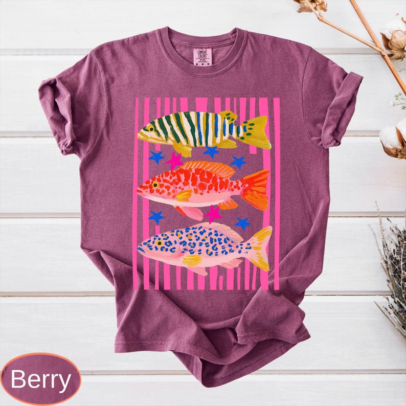 May include: Berry-colored t-shirt featuring a graphic of three colorful fish within a pink striped frame. The fish are yellow, orange, and blue, with star accents. The word "Berry" is at the bottom.