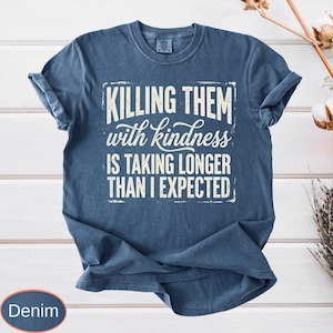 Op de afbeelding: Een denimblauw t-shirt met korte mouwen en een verweerde witte grafische print met de tekst "KILLING THEM with kindness IS TAKING LONGER THAN I EXPECTED" in een combinatie van dikke blokletters en een cursief lettertype.