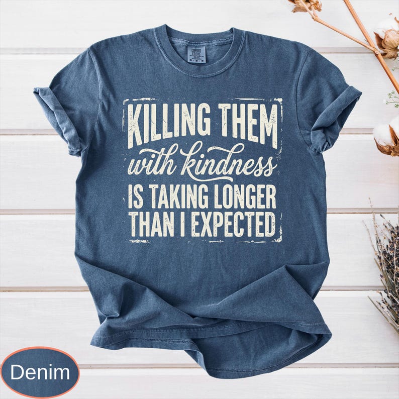 Op de afbeelding: Een denimblauw t-shirt met korte mouwen en een verweerde witte grafische print met de tekst "KILLING THEM with kindness IS TAKING LONGER THAN I EXPECTED" in een combinatie van dikke blokletters en een cursief lettertype.