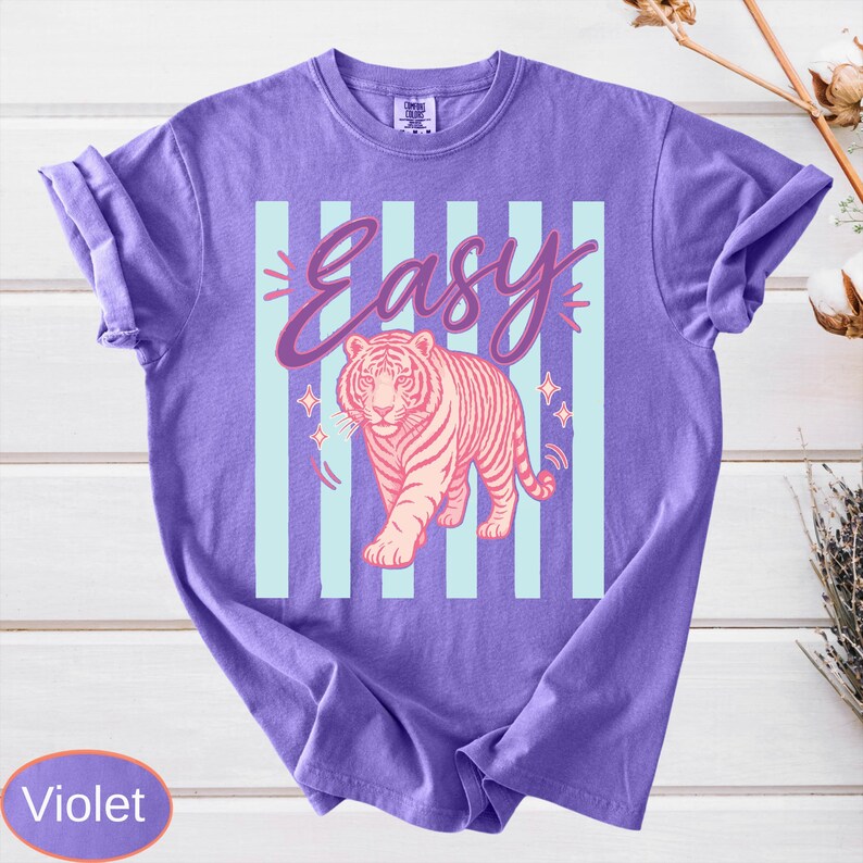 May include: Violet-colored t-shirt with a graphic design. The design features the word "Easy" in a cursive font above a pink tiger walking forward. Light blue vertical stripes and star accents complete the design.