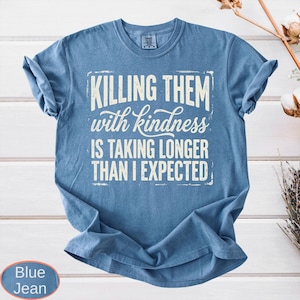 Op de afbeelding: Een jeansblauw t-shirt met korte mouwen en de witte tekst "KILLING THEM with kindness IS TAKING LONGER THAN I EXPECTED" gedrukt in een combinatie van blokletters en schrijfletters binnen een rechthoekige rand op de voorzijde.