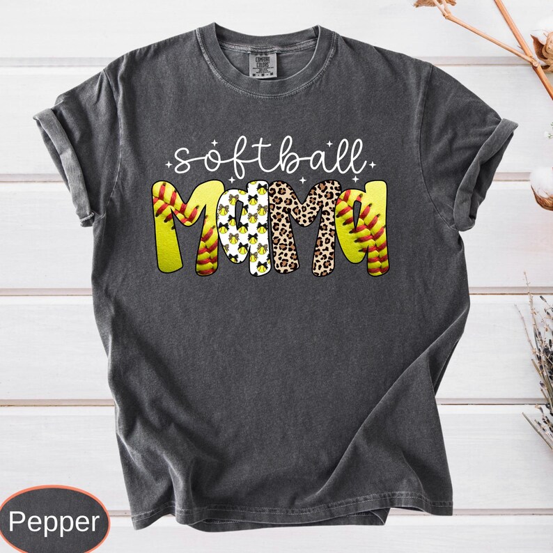 A vintage wash garment showing the text softball in cursive over the word MAMA. The thick letters showcase different designs like sports ball stitching, spotted animal print, and repeated bows, complete with tiny decorative sparkles.
