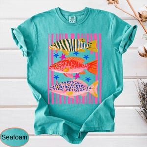May include: Seafoam green t-shirt featuring a colorful graphic of three fish. The fish are in yellow, orange, and pink, with blue and pink stars. The design is set against pink vertical stripes.