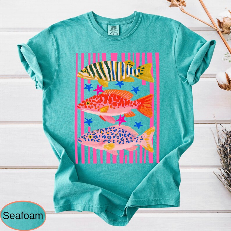 May include: Seafoam green t-shirt featuring a colorful graphic of three fish. The fish are in yellow, orange, and pink, with blue and pink stars. The design is set against pink vertical stripes.