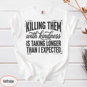 Op de afbeelding: Een wit t-shirt met korte mouwen en zwarte, verweerde tekst die luidt: KILLING THEM with kindness IS TAKING LONGER THAN I EXPECTED. De afbeelding is gecentreerd op de borst van het t-shirt, dat een klassieke ronde hals en korte mouwen heeft.