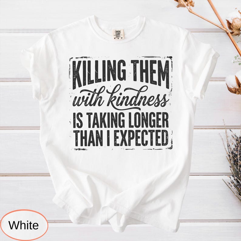 Op de afbeelding: Een wit t-shirt met korte mouwen en zwarte, verweerde tekst die luidt: KILLING THEM with kindness IS TAKING LONGER THAN I EXPECTED. De afbeelding is gecentreerd op de borst van het t-shirt, dat een klassieke ronde hals en korte mouwen heeft.