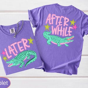 May include: Two violet t-shirts with a graphic of a green alligator. One shirt says "LATER" with pink text and a yellow star. The other says "AFTER WHILE" with pink text and yellow and pink stars.