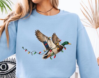 May include: A light blue crewneck sweatshirt featuring a graphic of a mallard duck in flight, wrapped in a string of colorful red, green, and yellow Christmas lights. The duck has a green head, brown chest, and patterned wings.