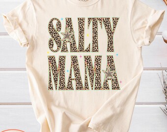 May include: An ivory short-sleeve t-shirt featuring the text SALTY MAMA in large block letters with a brown and black leopard print pattern. The design includes two small leopard print stars and several tiny colorful star icons.