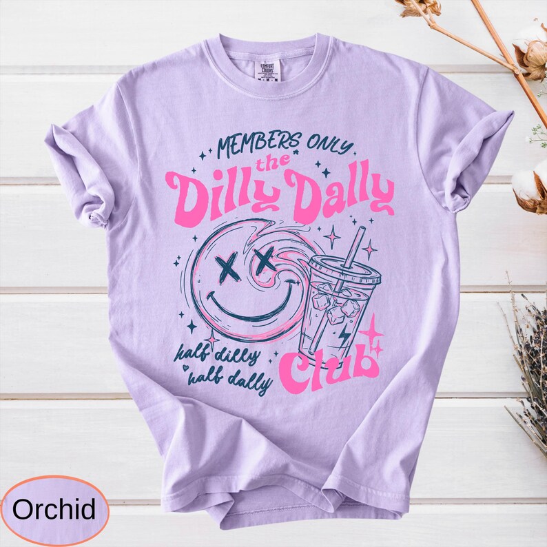 May include: Orchid-colored t-shirt with a graphic design. The design features a smiley face with an X for eyes, a drink, and the words "MEMBERS ONLY the Dilly Dally Club." The phrase "half dilly, half dally" is also included.