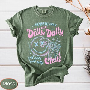 May include: Moss green t-shirt with a graphic design. The design features the text "MEMBERS ONLY the Dilly Dally Club" with a smiley face and a drink. The words "half dilly half dally" are also included.