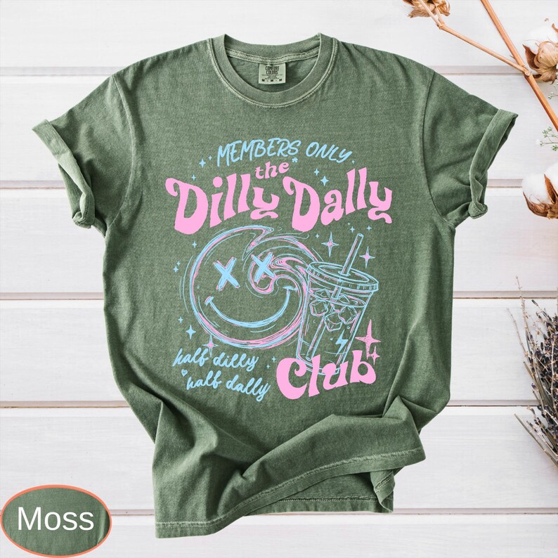 May include: Moss green t-shirt with a graphic design. The design features the text "MEMBERS ONLY the Dilly Dally Club" with a smiley face and a drink. The words "half dilly half dally" are also included.