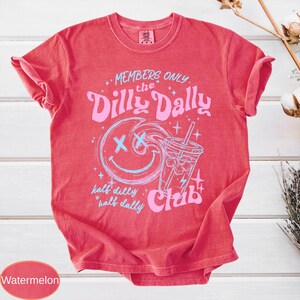 May include: Watermelon-colored t-shirt with a crew neck. The shirt features a graphic design with the text "MEMBERS ONLY the Dilly Dally Club" and a smiling face with an ice drink. The text "half dilly half dally" is also included.