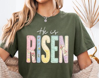 May include: Olive green t-shirt with the words "He is RISEN" in colorful, patchwork-style letters. The letters are in pink, blue, yellow, and floral patterns. The shirt has short sleeves and a crew neck.