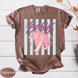 May include: Espresso-colored t-shirt featuring a pink tiger design with the word "Easy" in a purple script. The tiger is set against a backdrop of light blue vertical stripes, with star accents.
