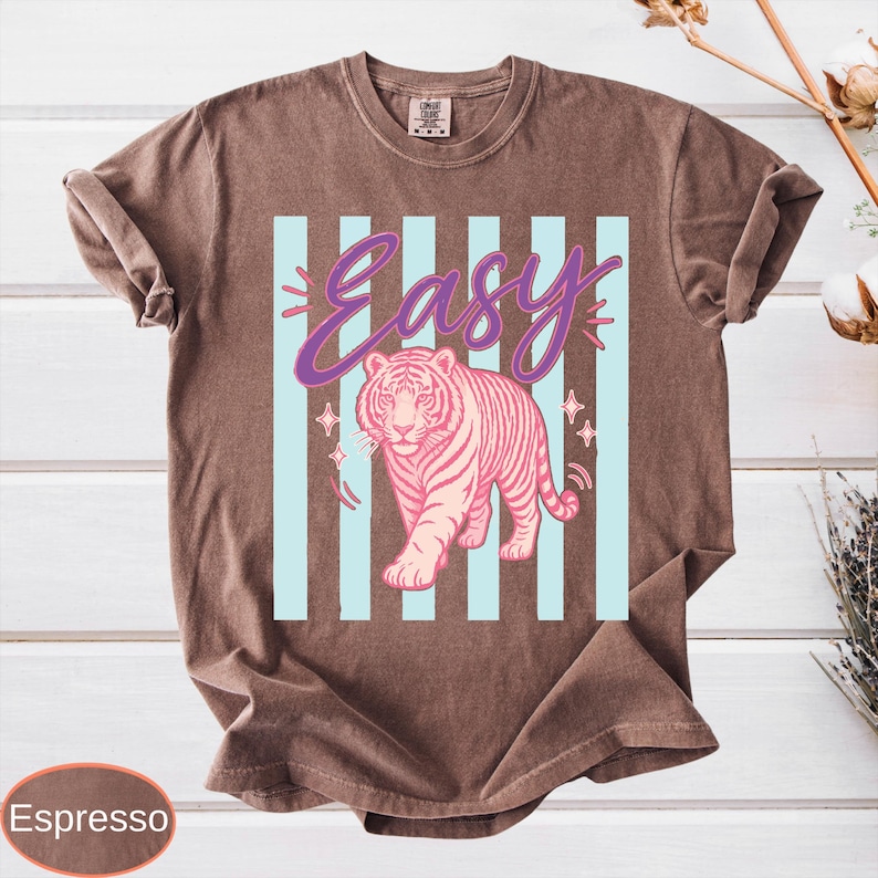 May include: Espresso-colored t-shirt featuring a pink tiger design with the word "Easy" in a purple script. The tiger is set against a backdrop of light blue vertical stripes, with star accents.