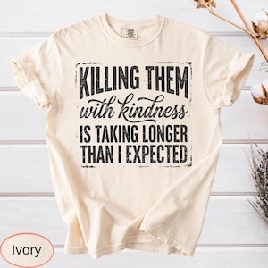 Op de afbeelding: Een ivoorkleurig T-shirt met korte mouwen en zwarte tekst: KILLING THEM with kindness IS TAKING LONGER THAN I EXPECTED. Het ontwerp is gecentreerd op de borst met een verweerde, rechthoekige rand rondom de dikgedrukte en cursieve letters.