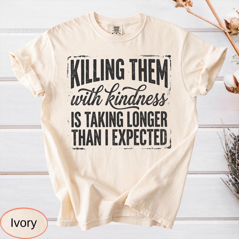 Op de afbeelding: Een ivoorkleurig T-shirt met korte mouwen en zwarte tekst: KILLING THEM with kindness IS TAKING LONGER THAN I EXPECTED. Het ontwerp is gecentreerd op de borst met een verweerde, rechthoekige rand rondom de dikgedrukte en cursieve letters.