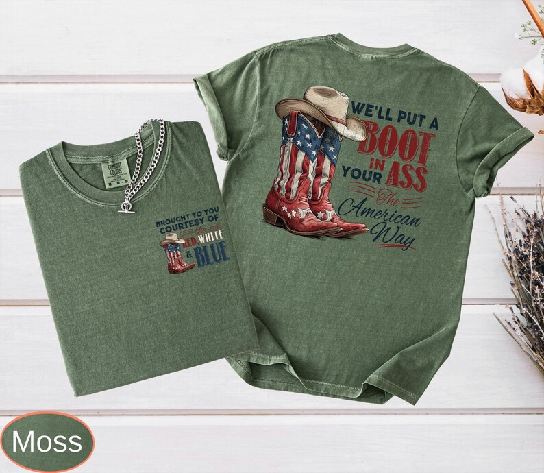 May include: Two moss green t-shirts with graphic designs. One shirt features a cowboy boot with the American flag and the text "BROUGHT TO YOU COURTESY OF WHITE & BLUE". The other shirt has a cowboy boot with the American flag and the text "WE'LL PUT A BOOT IN YOUR ASS The American Way".