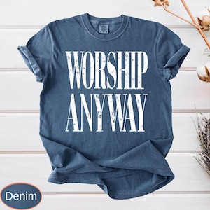 May include: Denim blue t-shirt with the words "WORSHIP ANYWAY" in large white letters. The shirt has short sleeves and a crew neck. The shirt is displayed on a white wooden surface.