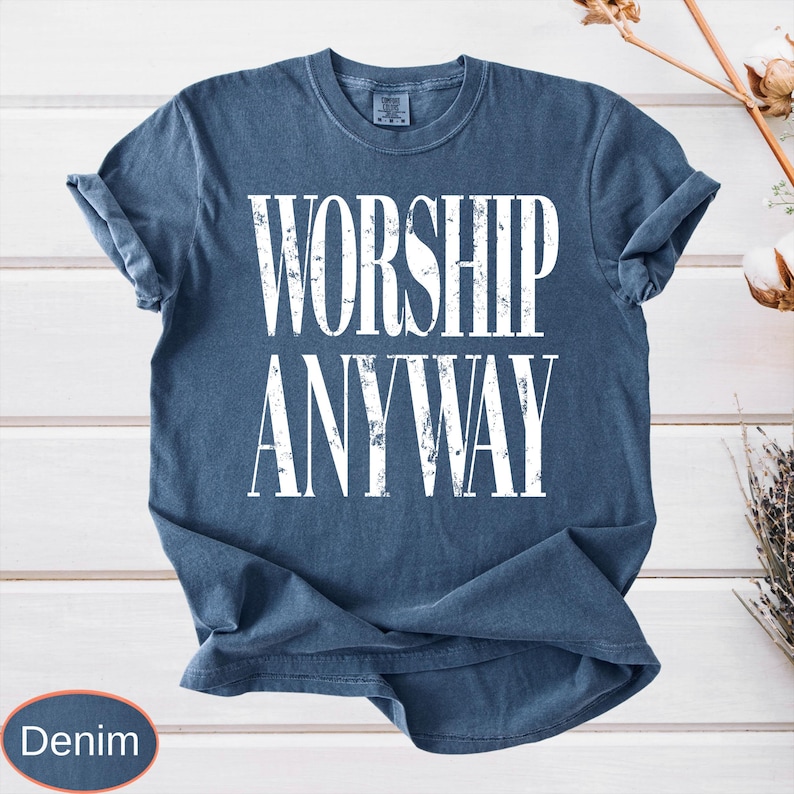 May include: Denim blue t-shirt with the words "WORSHIP ANYWAY" in large white letters. The shirt has short sleeves and a crew neck. The shirt is displayed on a white wooden surface.