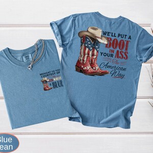 May include: Two blue jean t-shirts. One shirt has a small graphic of a cowboy boot with the text "BROUGHT TO YOU COURTESY OF RED, WHITE & BLUE". The other shirt has a graphic of a cowboy boot with the American flag design and the text "WE'LL PUT A BOOT IN YOUR ASS The American Way".