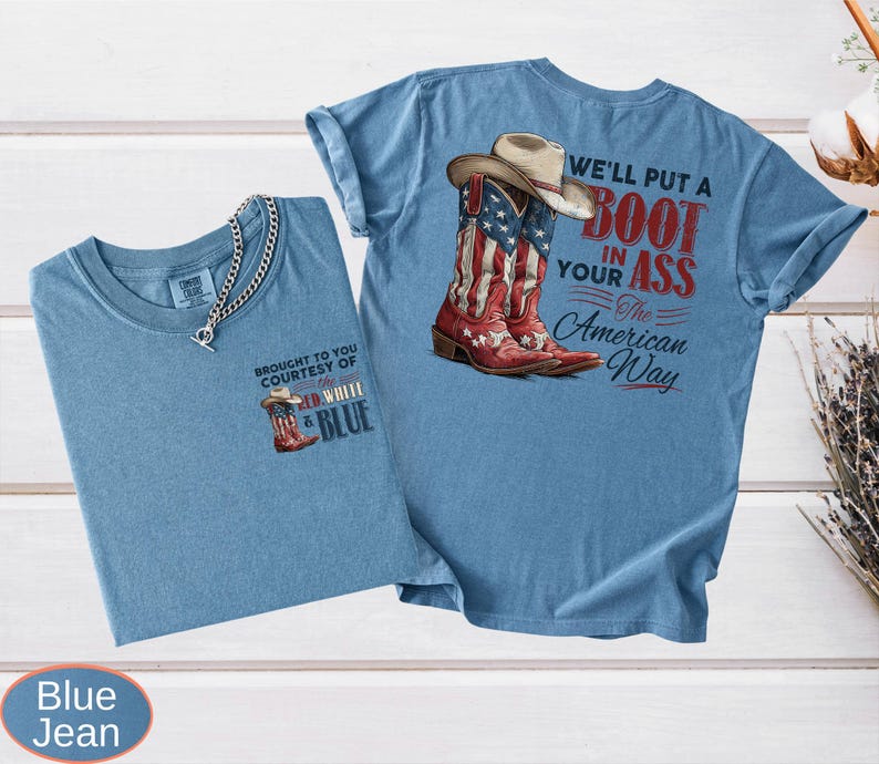 May include: Two blue jean t-shirts. One shirt has a small graphic of a cowboy boot with the text "BROUGHT TO YOU COURTESY OF RED, WHITE & BLUE". The other shirt has a graphic of a cowboy boot with the American flag design and the text "WE'LL PUT A BOOT IN YOUR ASS The American Way".