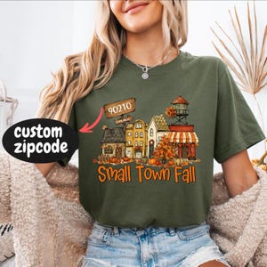 May include: An olive green short-sleeve t-shirt featuring a graphic of a small town street with autumn trees, pumpkins, and a water tower. A wooden sign displays the text 90210, and the text Small Town Fall is printed in orange below the buildings.